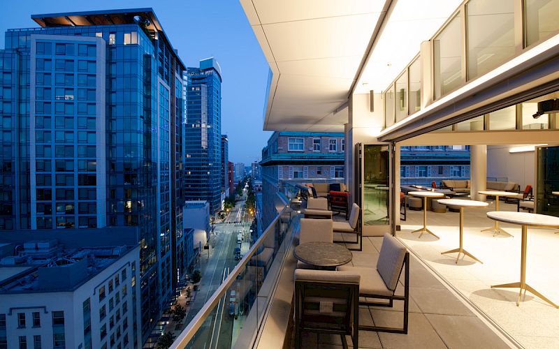 A modern urban balcony with chairs and tables overlooks a city street and high-rise buildings at dusk, with blue sky and street lights on.