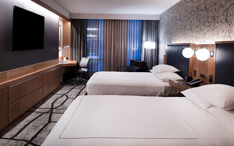 A modern hotel room with two beds, a TV, a desk, and lamps, characterized by stylish decor and soft lighting, creates a comfortable atmosphere.