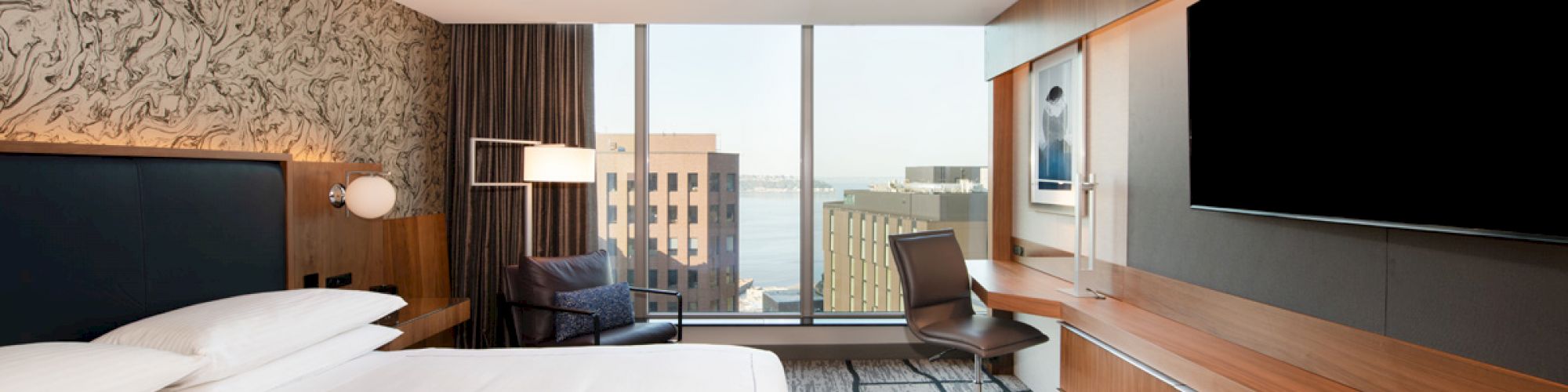 A modern hotel room with a large bed, a desk with a chair, a large TV, and a window offering a city view.