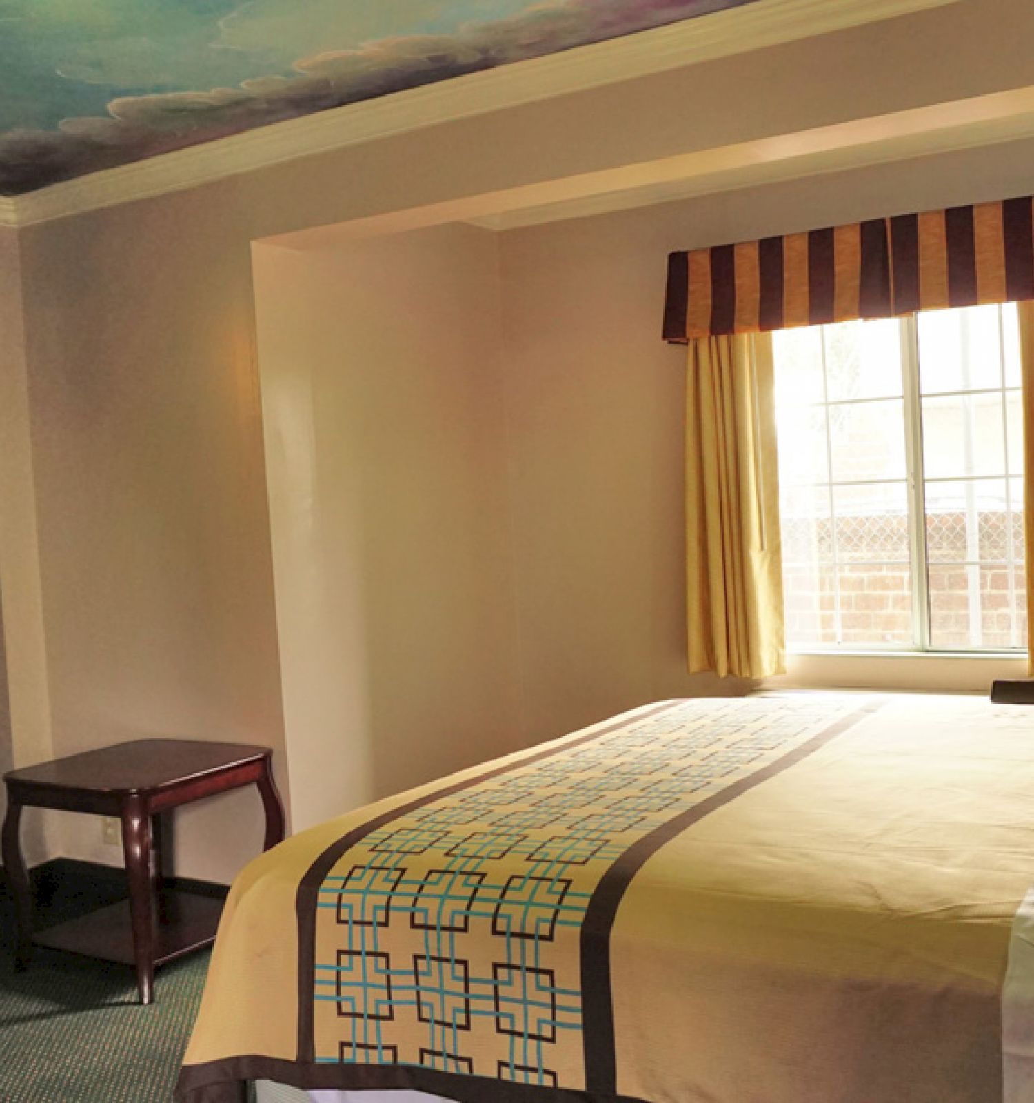 A hotel room with a bed, side table, wall-mounted TV, window with curtains, and a painted sky ceiling.