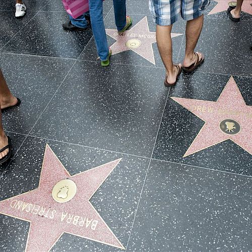 People are walking on the Hollywood Walk of Fame, with visible stars for Barbara Stanwyck and Drew Barrymore.