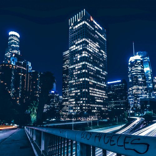 A nighttime urban scene featuring illuminated skyscrapers, a busy highway with light trails, and a railing with 