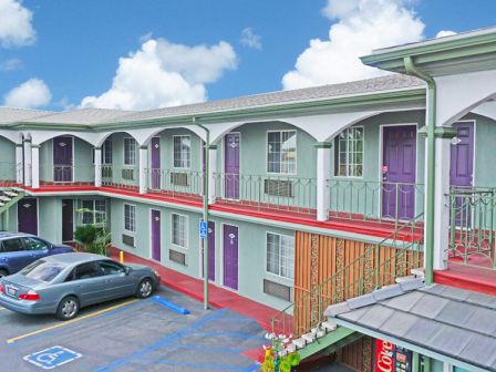 A two-story motel with purple doors, outdoor stairs, and parked cars in the lot beneath a blue sky with clouds.