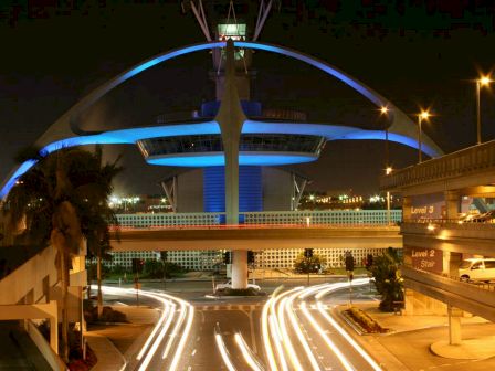 The image shows the illuminated Theme Building at Los Angeles International Airport at night, with light trails of vehicles passing by.