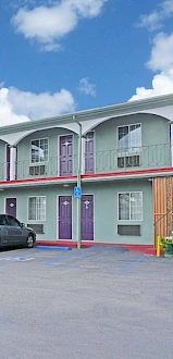 A two-story motel with parked cars, flower pots, and bright weather, featuring green walls, purple doors, and external staircases.